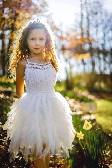 Young girl with curly hair standing outdoors in a white dress with a fluffy skirt, surrounded by trees with autumn foliage and flowers.