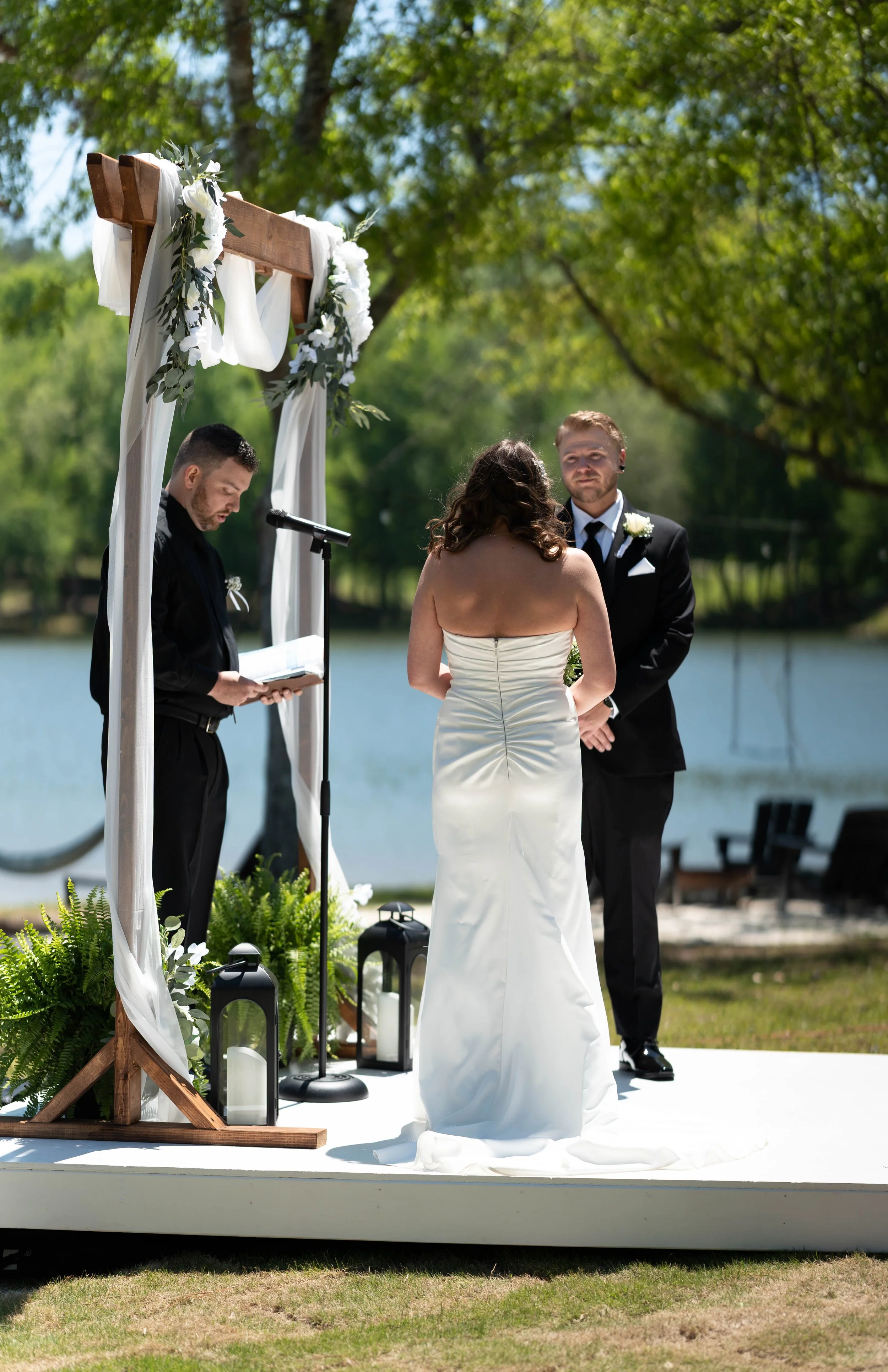 A bride and groom exchanging vows outdoors by the water during a wedding ceremony, with a officiant reading from a book under a wooden arch decorated with white fabric and flowers.