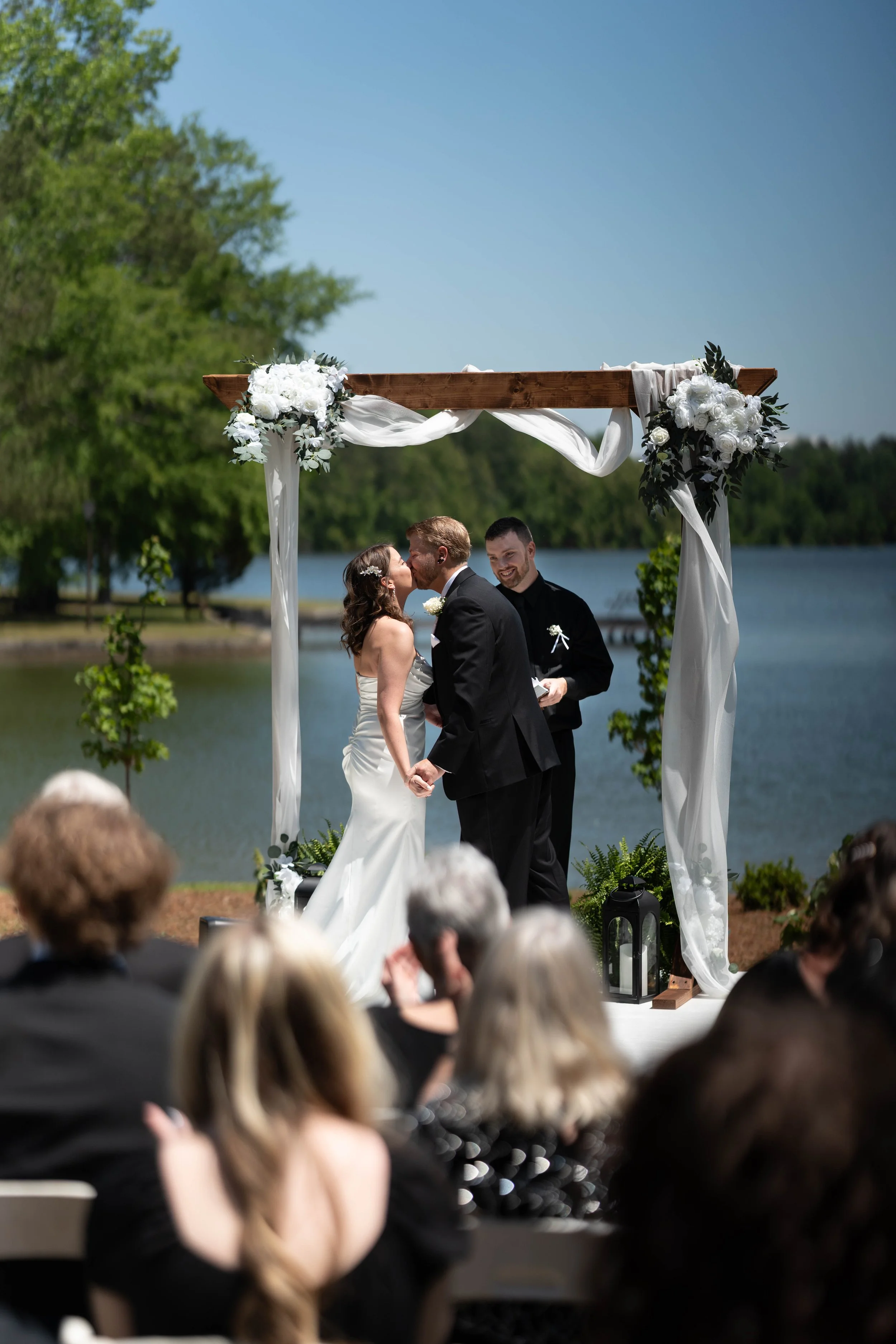 A couple getting married outdoors by a lake, kissing under a wooden arch decorated with white flowers and drapery, with a officiant standing nearby, guests seated in the foreground, and trees in the background.