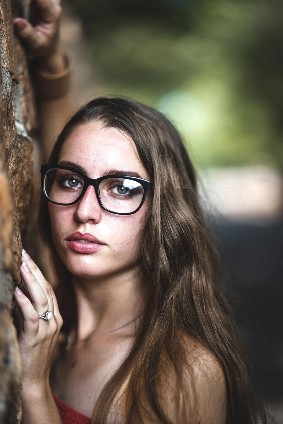 A young woman with long brown hair and black glasses is leaning against a brick wall. She has light skin and is wearing a red sleeveless top. Her left hand, with a ring on her finger, is resting on the wall near her face. The background is blurred with hints of green and light, suggesting an outdoor setting.