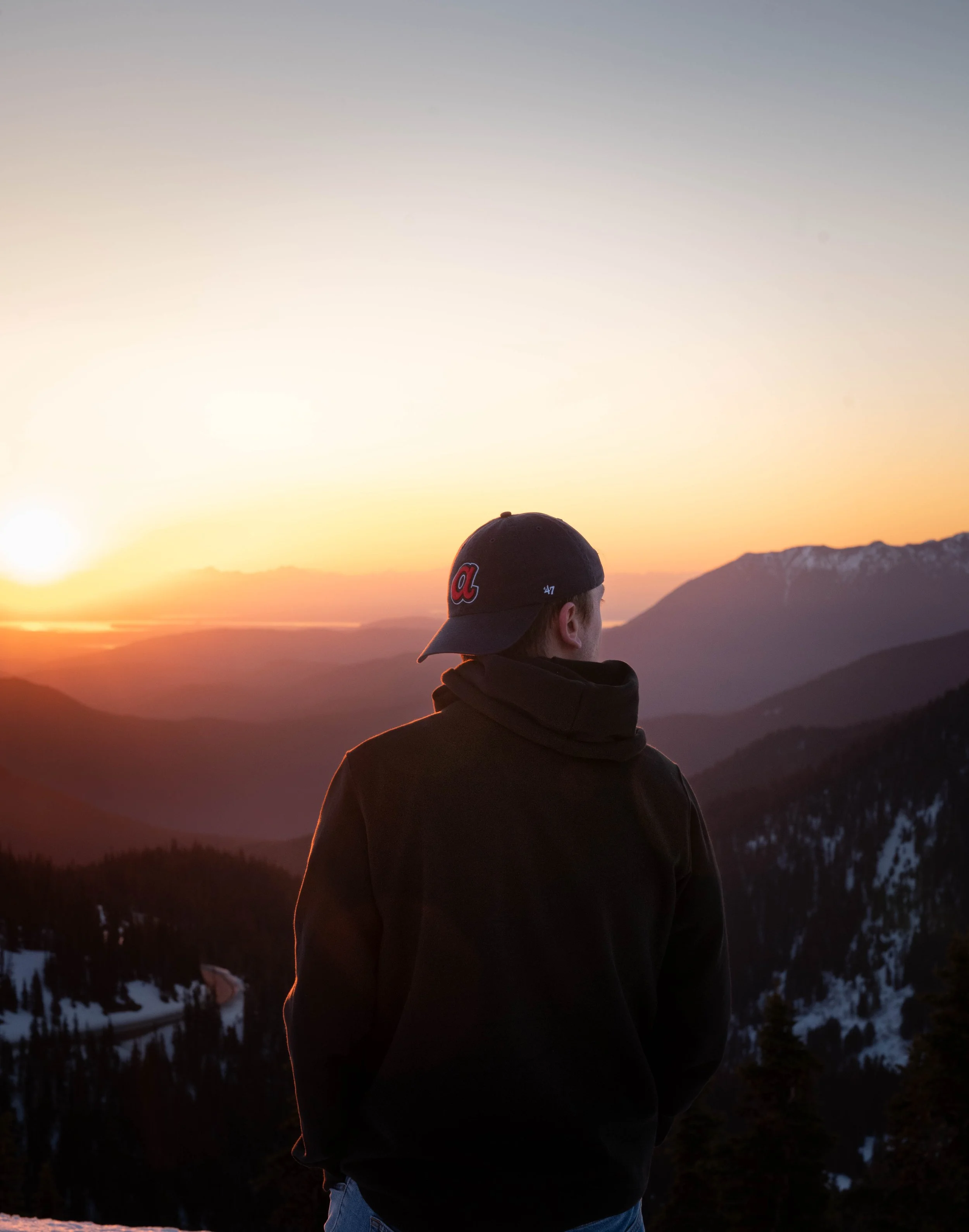 A person wearing a black jacket and baseball cap standing outdoors during sunset, overlooking a mountain landscape with snow patches and trees.