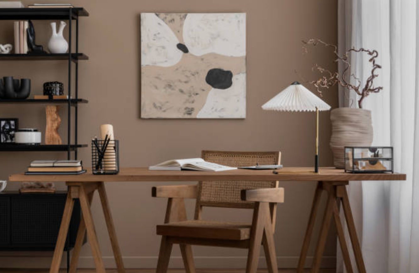 A home office with a wooden desk, a chair with a woven back, a black wire pen holder, an open book, decorative items, a table lamp, a large vase with dried branches, a framed photo, a bookshelf, a painting on the wall, and curtains on the window.