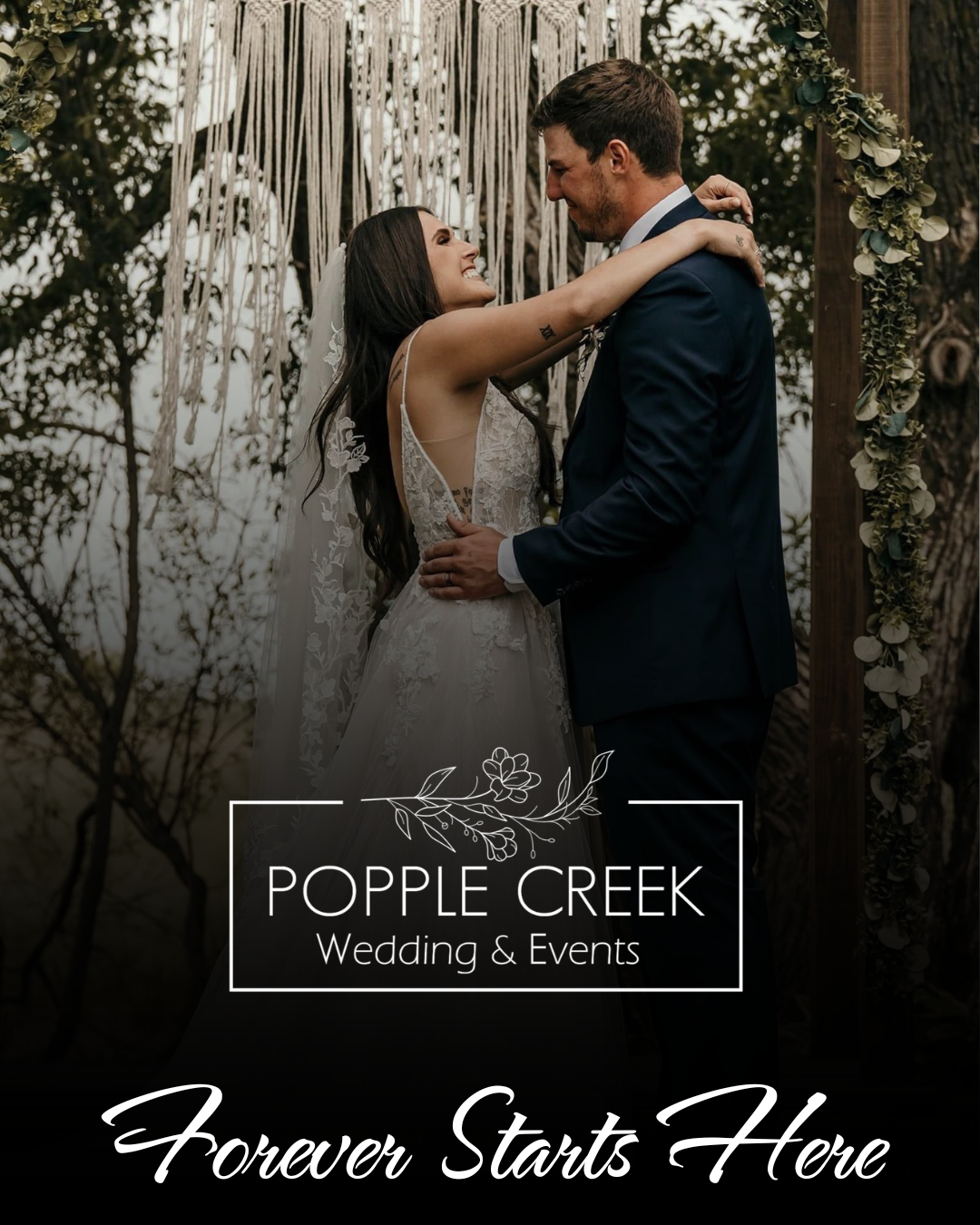 A bride and groom smiling and embracing during their outdoor wedding ceremony, standing in front of a decorated arch with greenery and fabric strands, surrounded by trees.