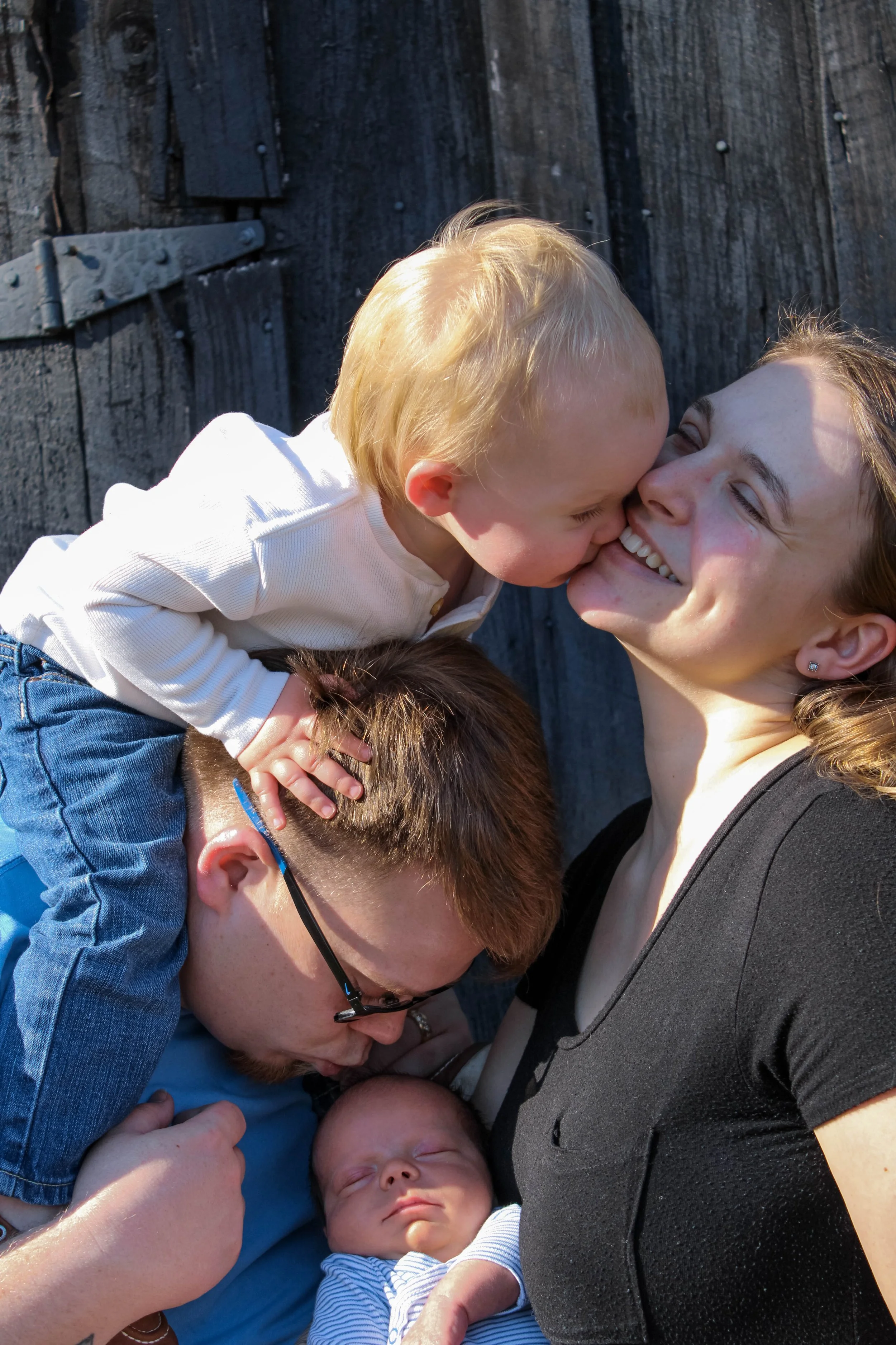 A young woman with brown hair smiling and looking at a blonde boy giving her a kiss on the nose, with a baby lying nearby asleep, all in front of a dark wooden fence.
