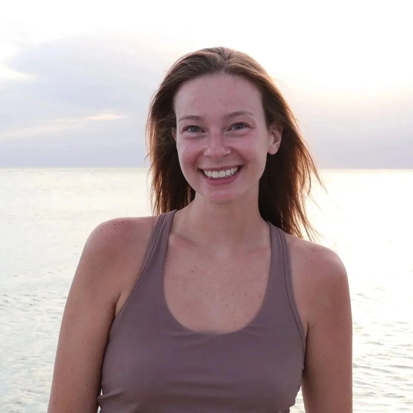 Smiling woman with reddish brown hair standing near a body of water during sunset, wearing a purple tank top.