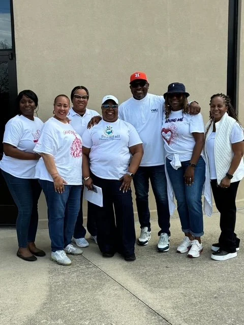Group of seven people standing outdoors against a beige wall, smiling, wearing casual clothing including white t-shirts, jeans, and sneakers.