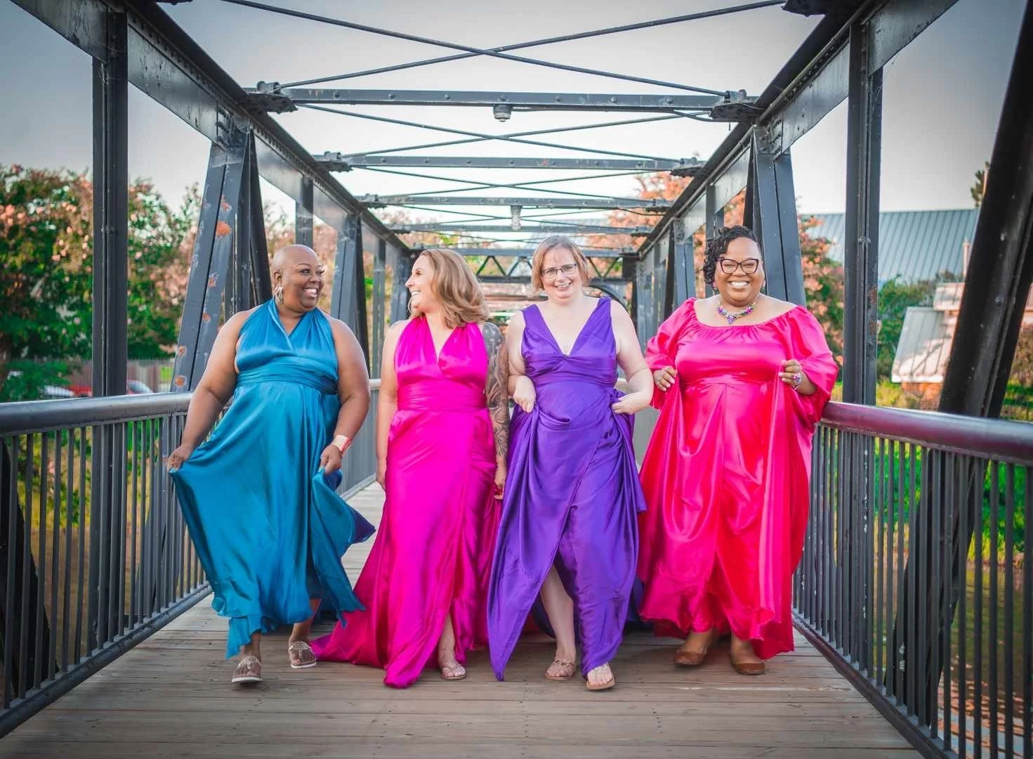 Four women in colorful evening gowns walking on a bridge, laughing and enjoying each other's company.