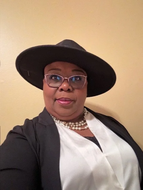 A woman wearing a black wide-brim hat, glasses, a pearl necklace, and a black blazer over a white blouse, standing against a beige wall.