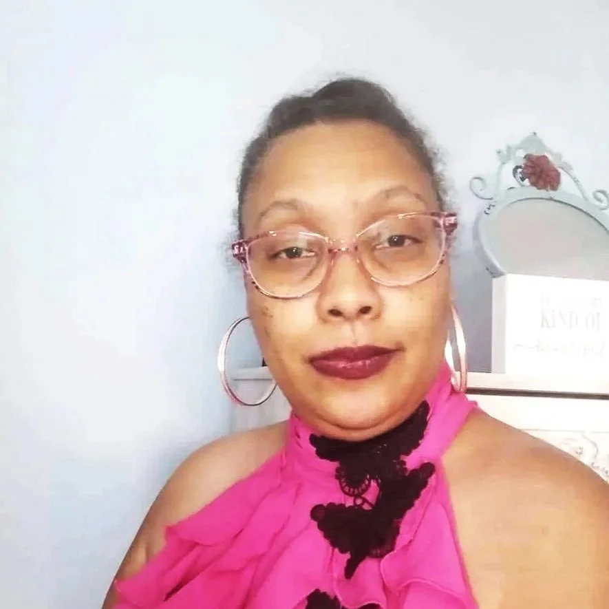 A woman with short hair, wearing glasses, large hoop earrings, dark lipstick, and a pink top with black lace details, standing in front of a light-colored wall and a decorative dresser.