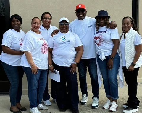 Group of seven diverse women and one man standing together outdoors, smiling, wearing casual clothes including white t-shirts with designs and logo, some wearing hats and glasses.