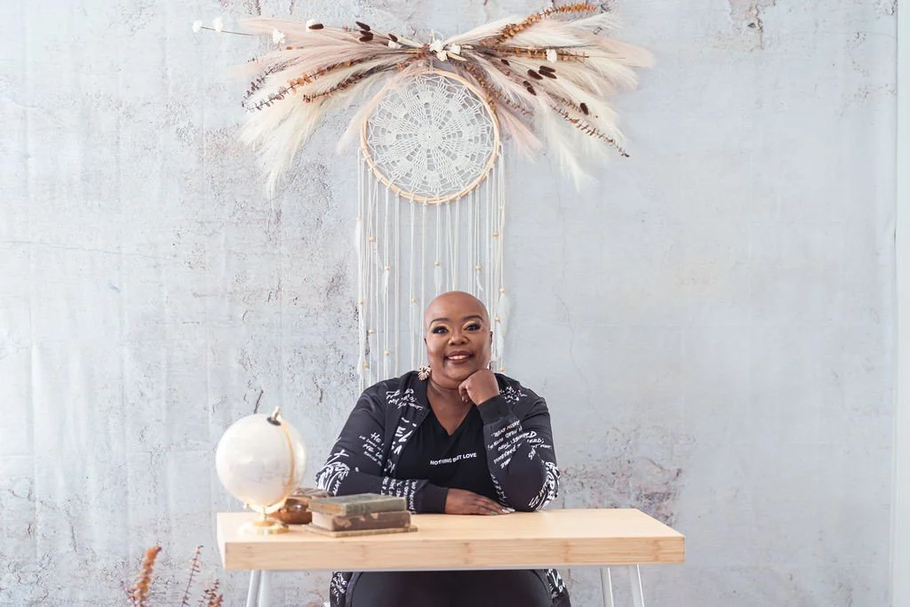 A woman sitting at a desk in front of a decorative wall hanging with feathers and a dreamcatcher.
