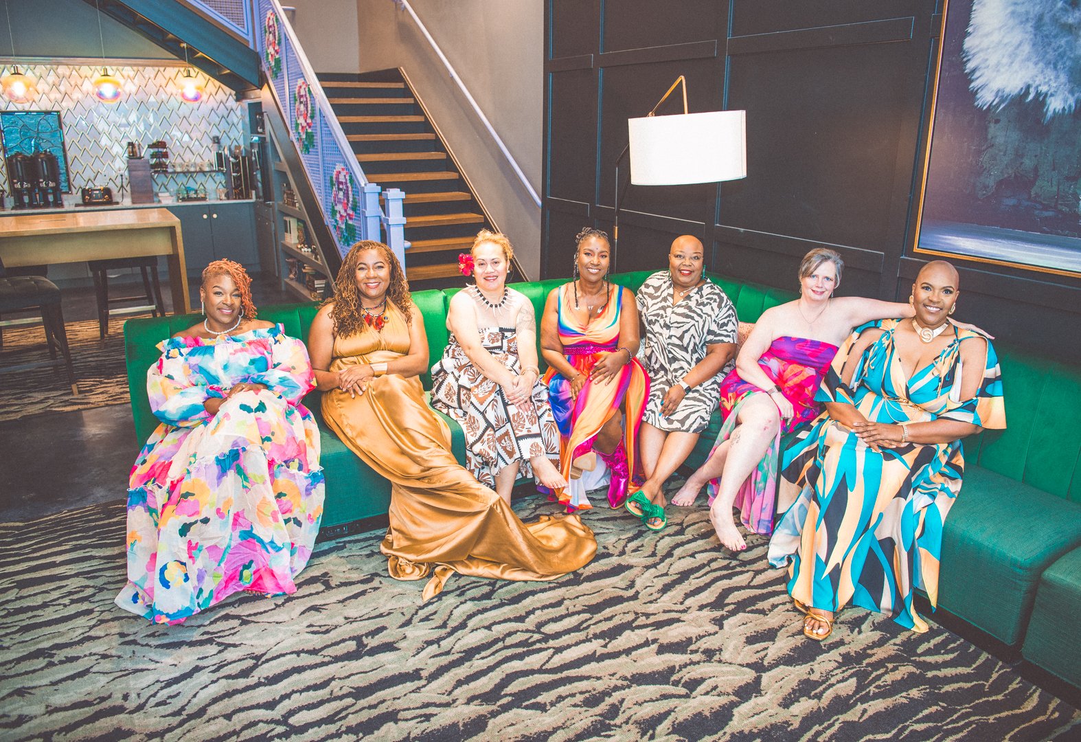 Seven women in colorful dresses sitting on a green couch in a hotel lobby or lounge area with a staircase and bar in the background.