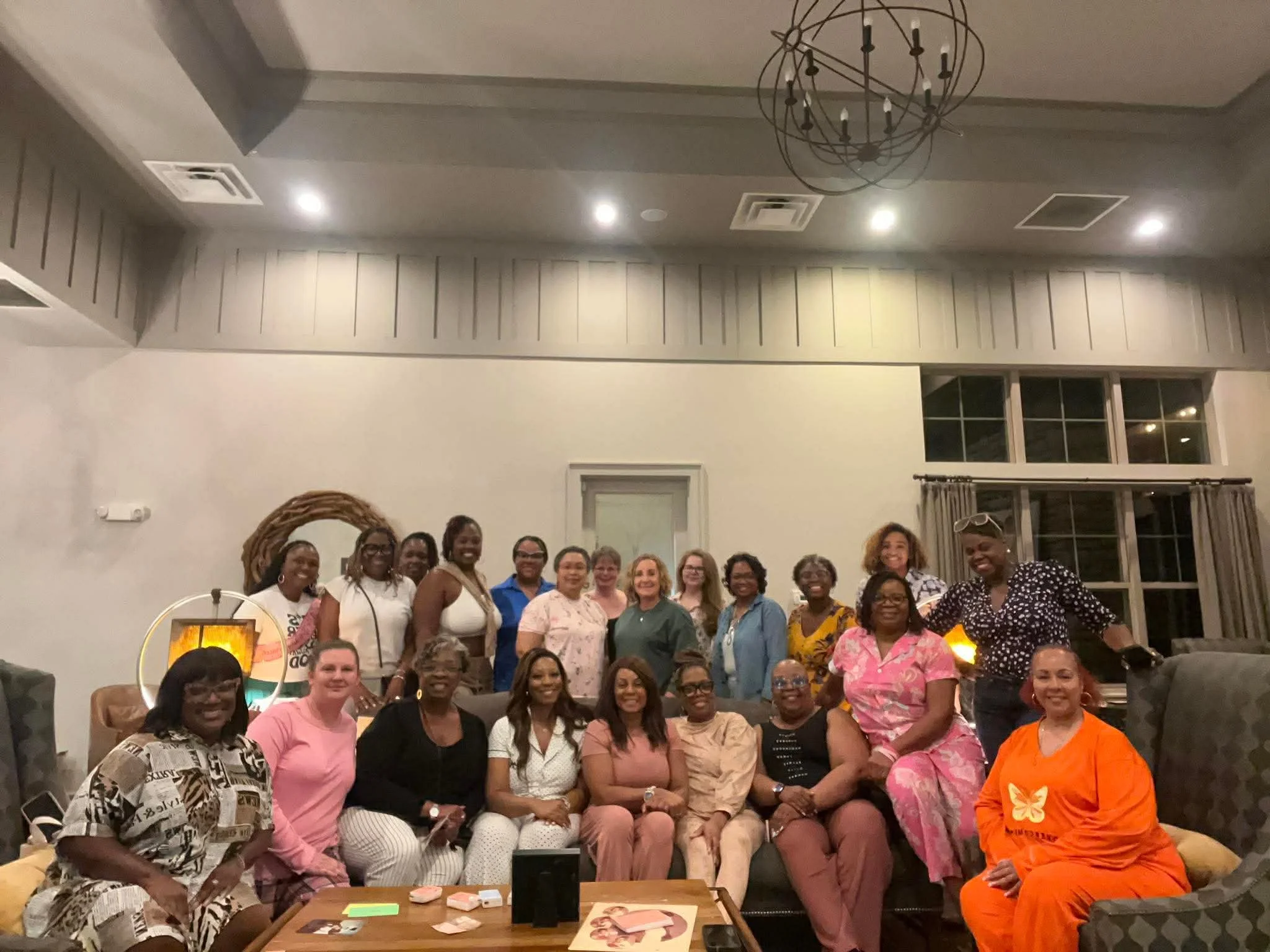 A large group of women gathered in a warmly lit room, smiling for a group photo. The room features a modern chandelier, a window with curtains, and a side table with orange candles and decorative items. The women are dressed in colorful and patterned clothing, sitting and standing close together.