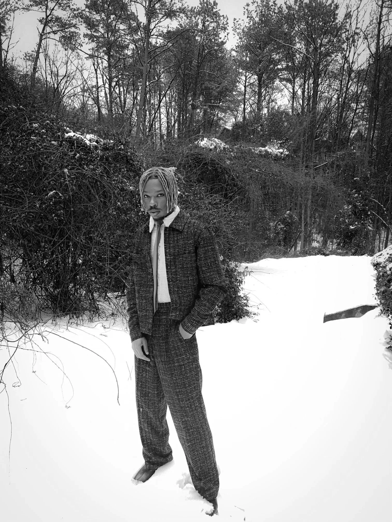 A person with dreadlocks wearing a plaid suit and boots standing in the snow in a winter wooded landscape.