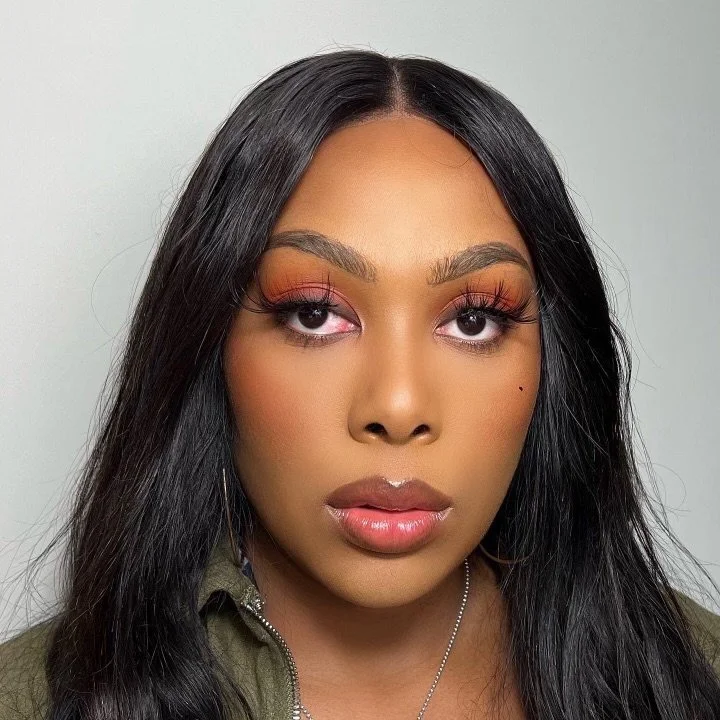 Close-up portrait of a young woman with long black hair and makeup featuring orange eyeshadow, pink blush, and glossy lips, wearing a green jacket and a silver necklace.