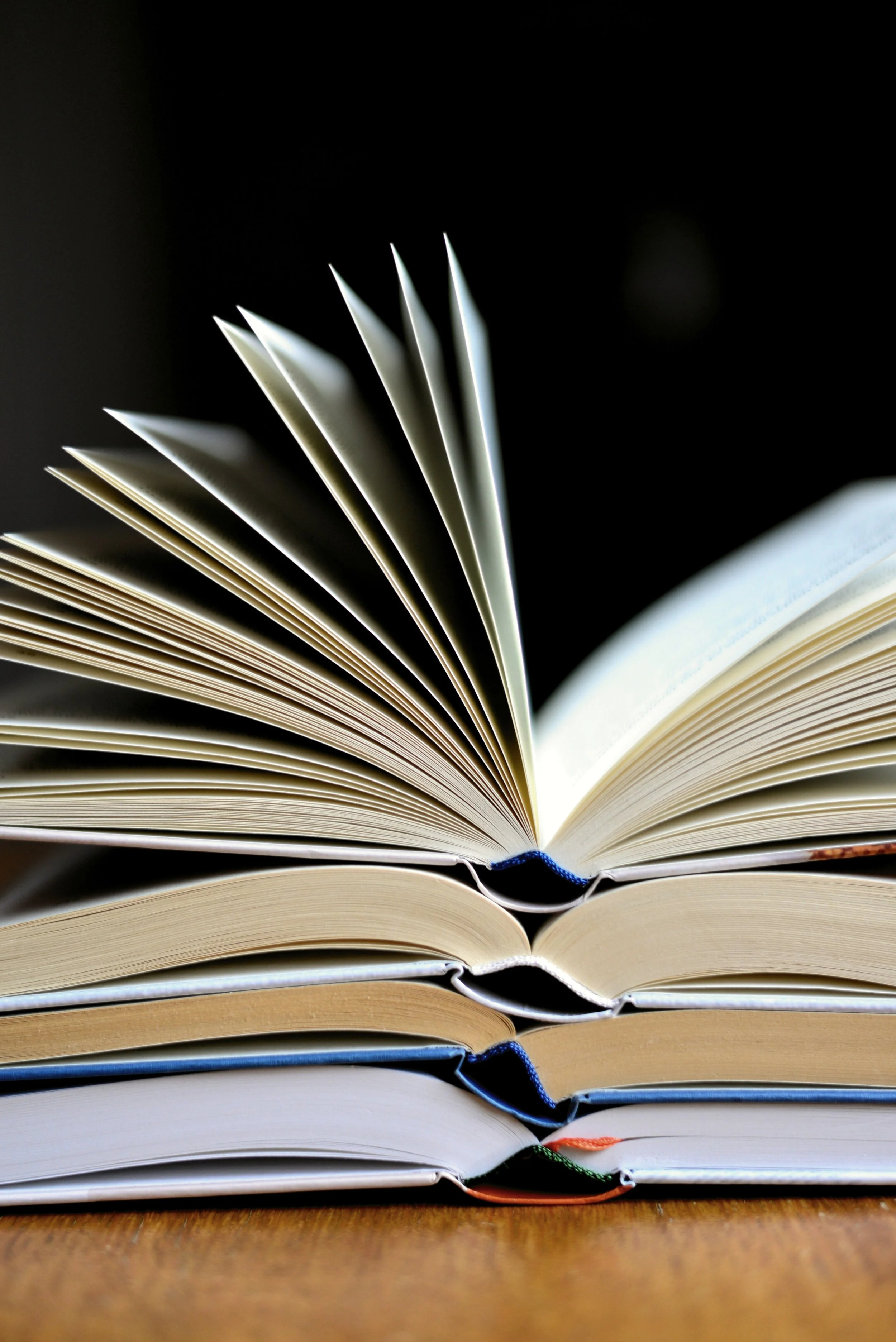 Stack of open books with pages fanned out on a wooden surface against a black background.
