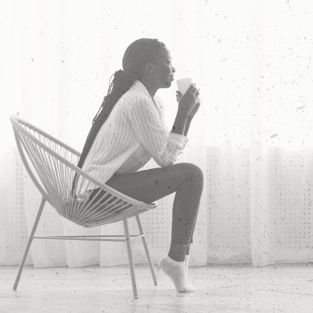 Woman sitting on a modern chair, holding a mug, looking out a window, wearing a striped shirt, pants, and socks, in bright natural light.