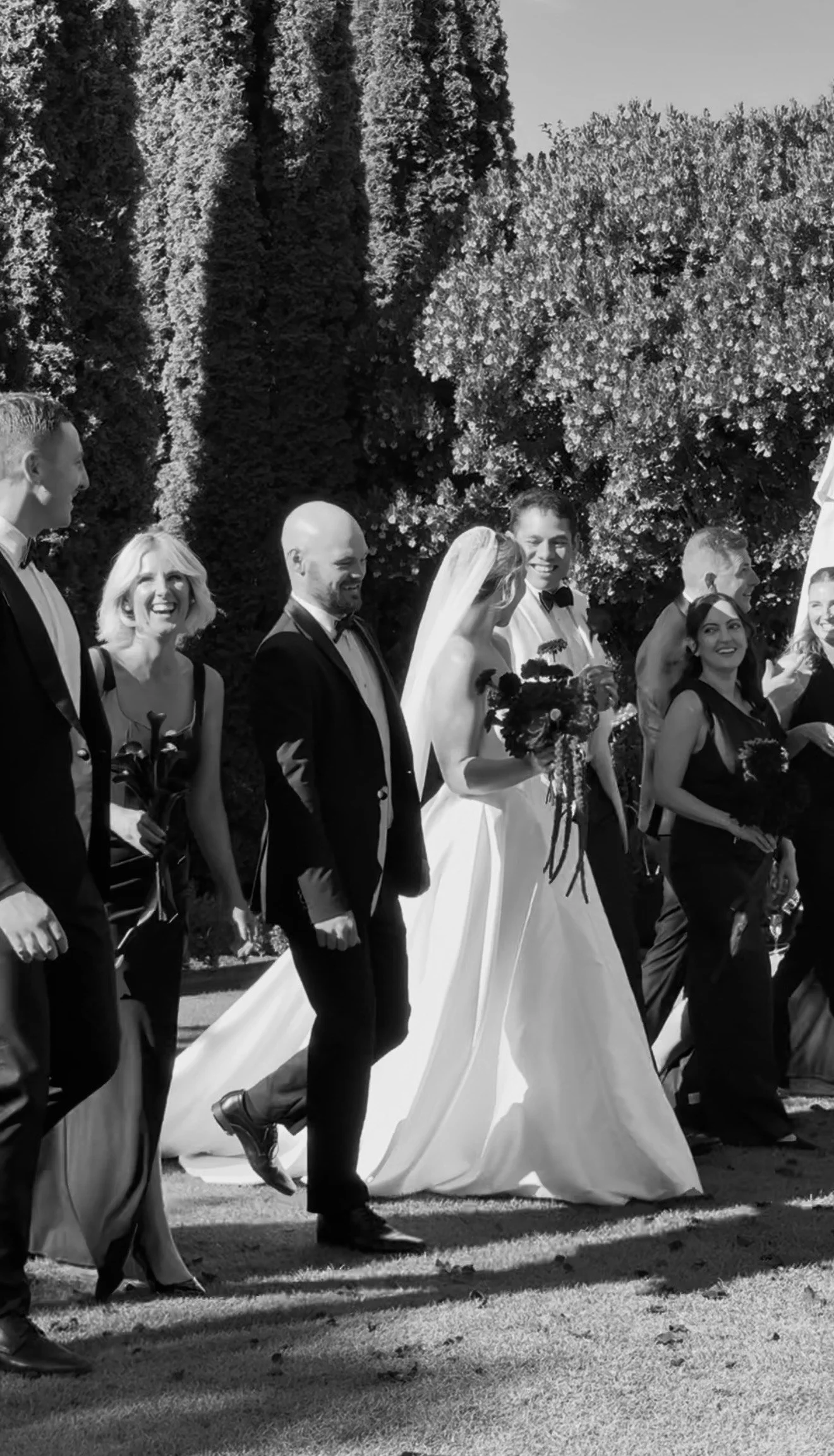 Black and white photo of a wedding party walking outdoors, with a bride in a gown and veil holding a bouquet, surrounded by men and women dressed in formal attire, and tall trees in the background.