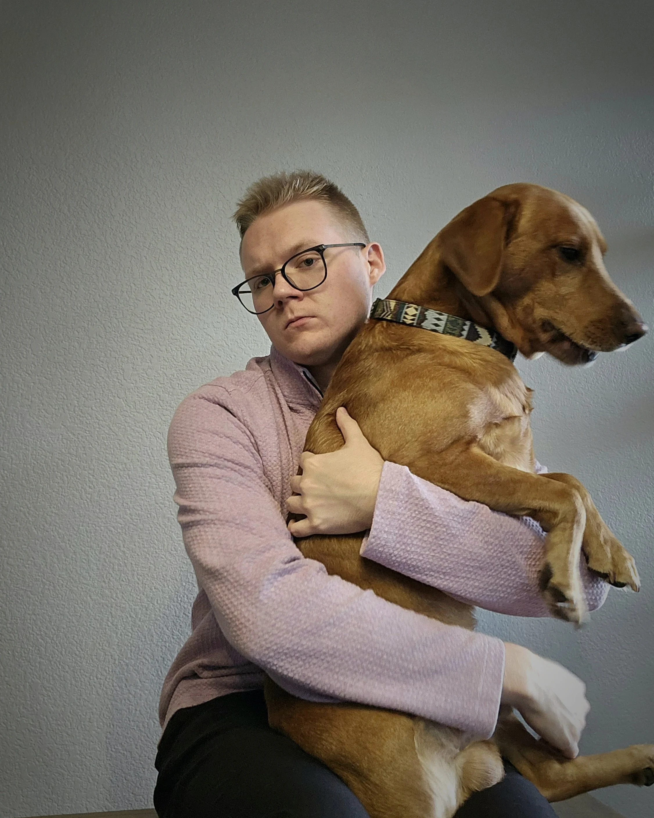 A young man with glasses and short hair is sitting on a bench, holding a large brown dog with a patterned collar. The dog is sitting on his lap, facing sideways, and the man is looking directly at the camera. The background is a plain, textured light gray wall.