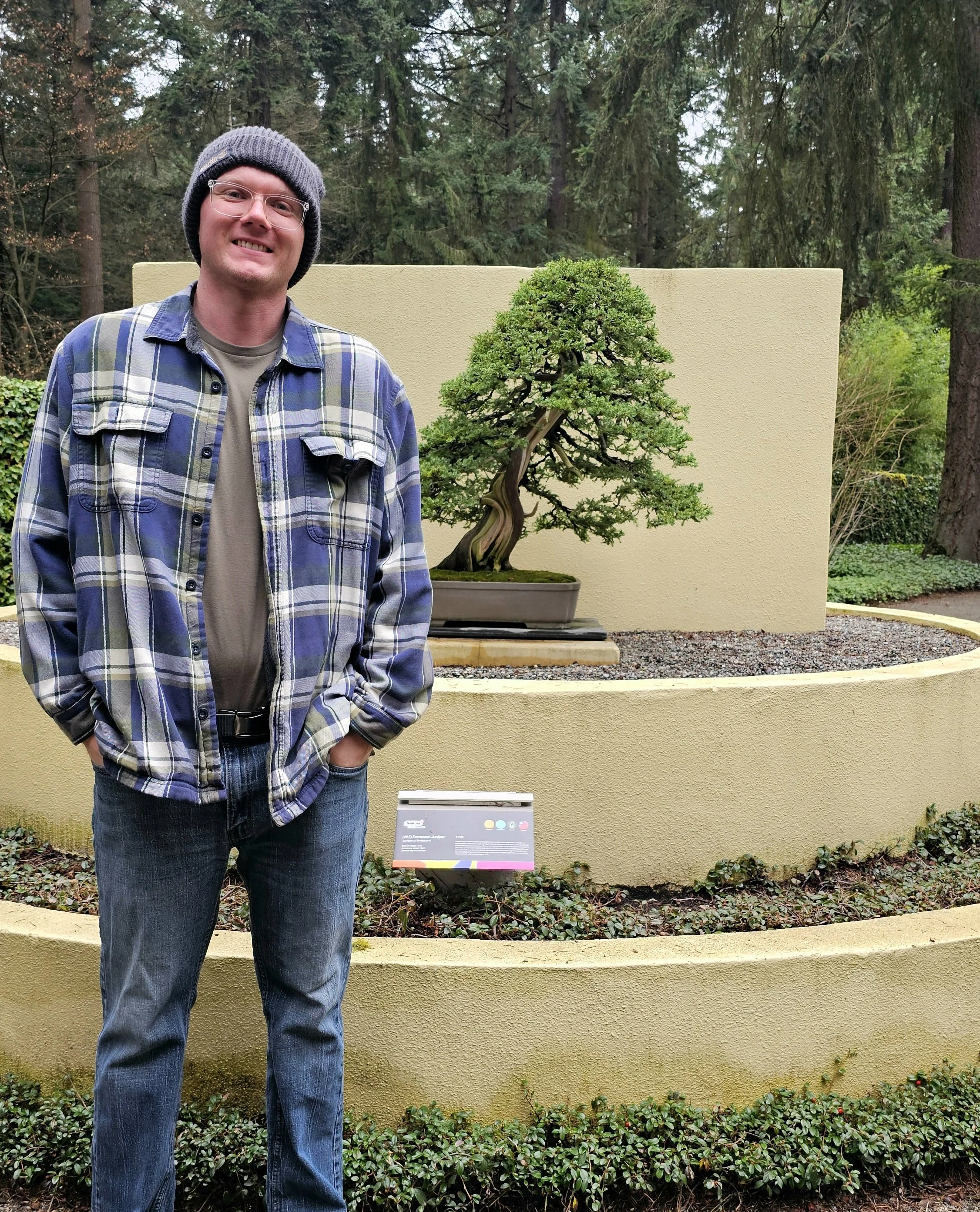 A man wearing glasses, a gray beanie, a plaid jacket, and jeans standing outdoors in front of a bonsai tree display with forest in the background.