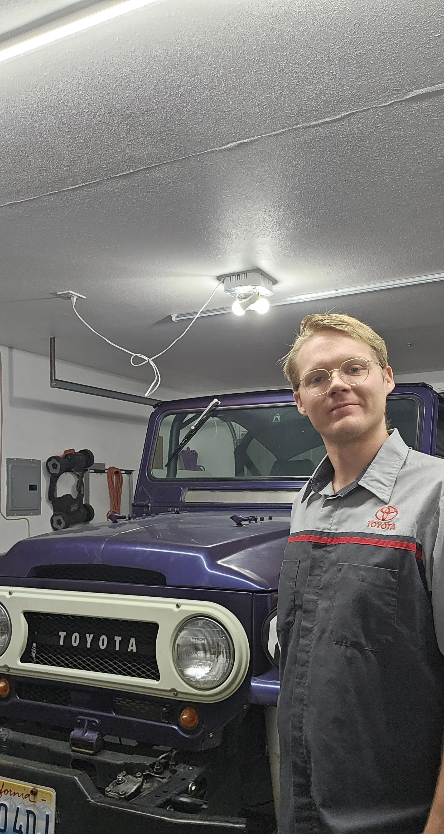 Young man wearing glasses and a Toyota mechanic uniform standing in garage behind purple Toyota SUV with open hood, car in repair.
