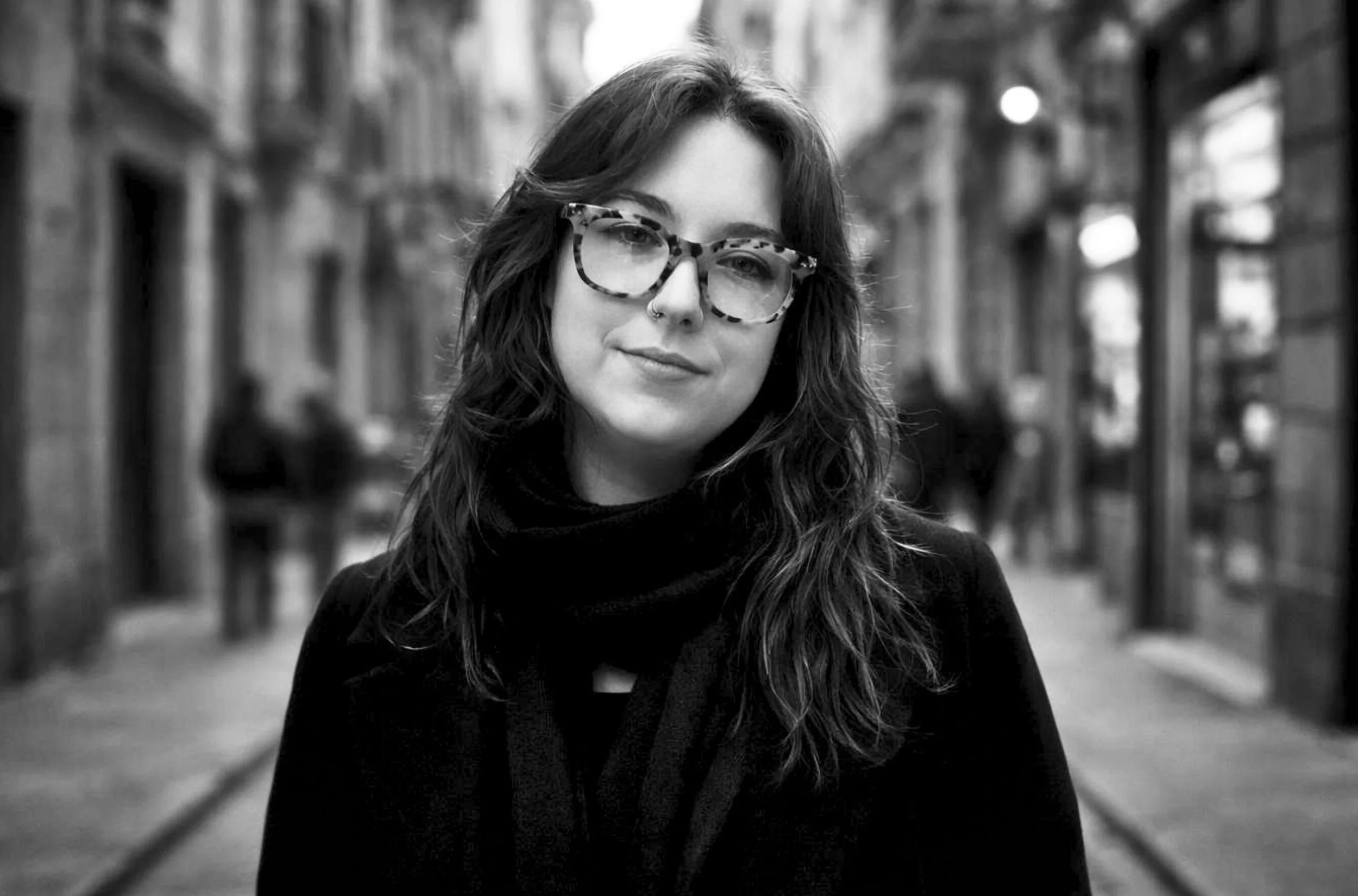 Black and white portrait of a young woman with wavy hair, wearing glasses and a nose piercing, standing outdoors in an alley with blurred buildings and lights in the background.