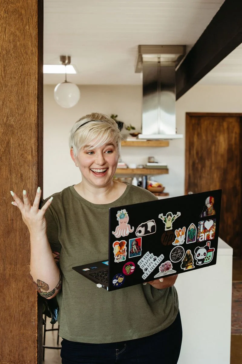 Lauren Abbott looking at a laptop decorated with various stickers, in a cozy kitchen setting.
