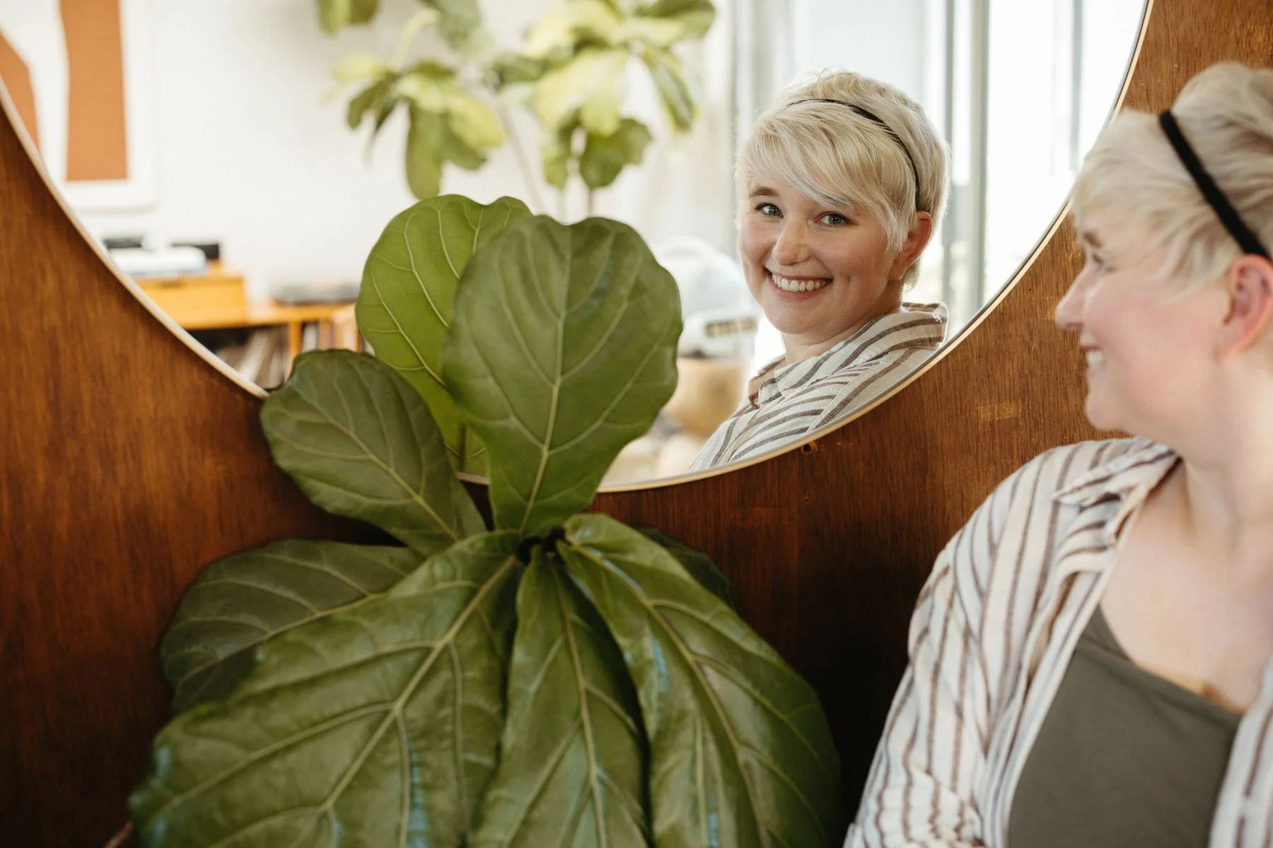 A woman with short blonde hair wearing a striped shirt looking in a circular mirror, smiling, reflection visible, green houseplant in foreground, indoor setting with natural light.