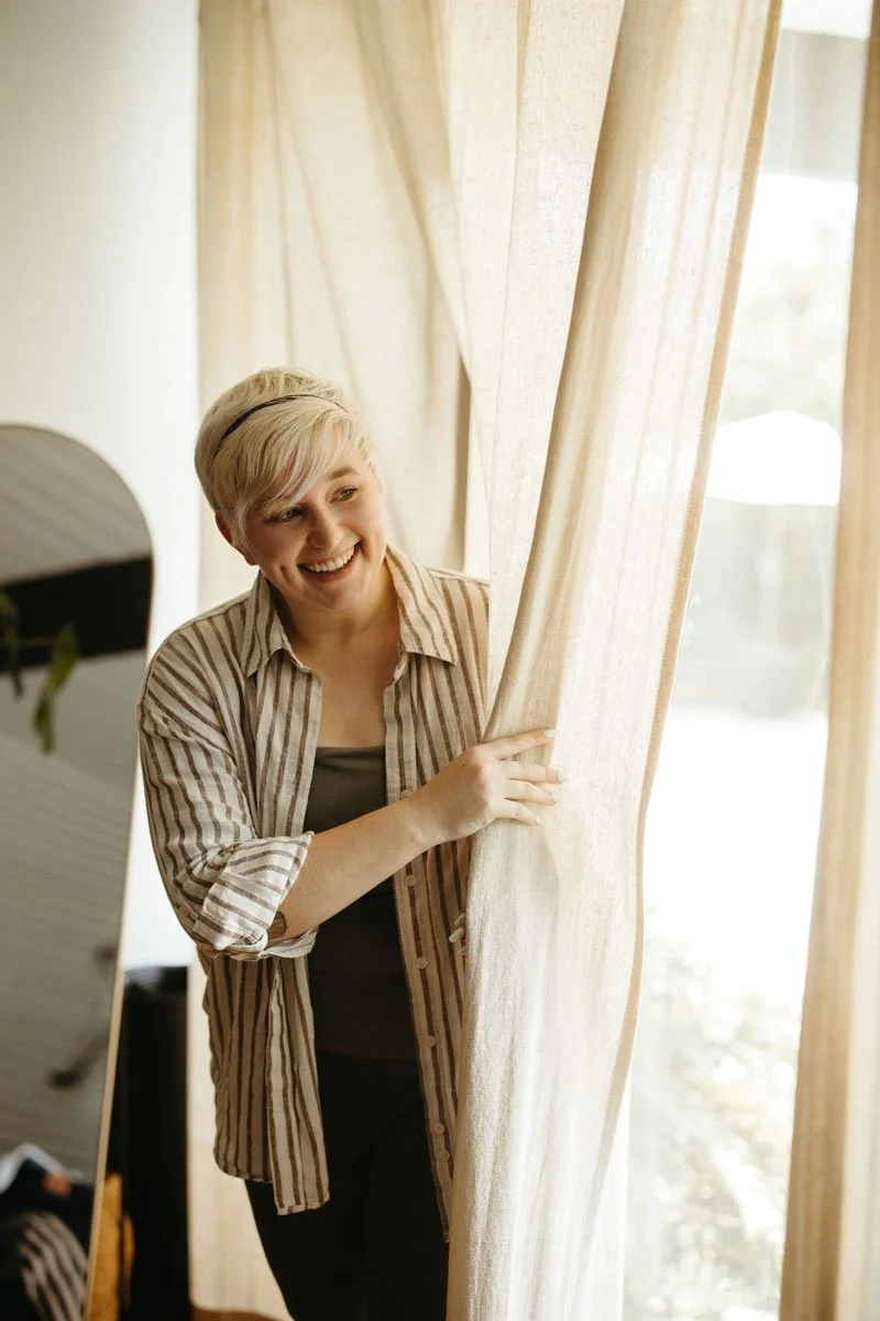 A woman with short blonde hair and a striped shirt smiling while peeking through beige curtains in a bright room.