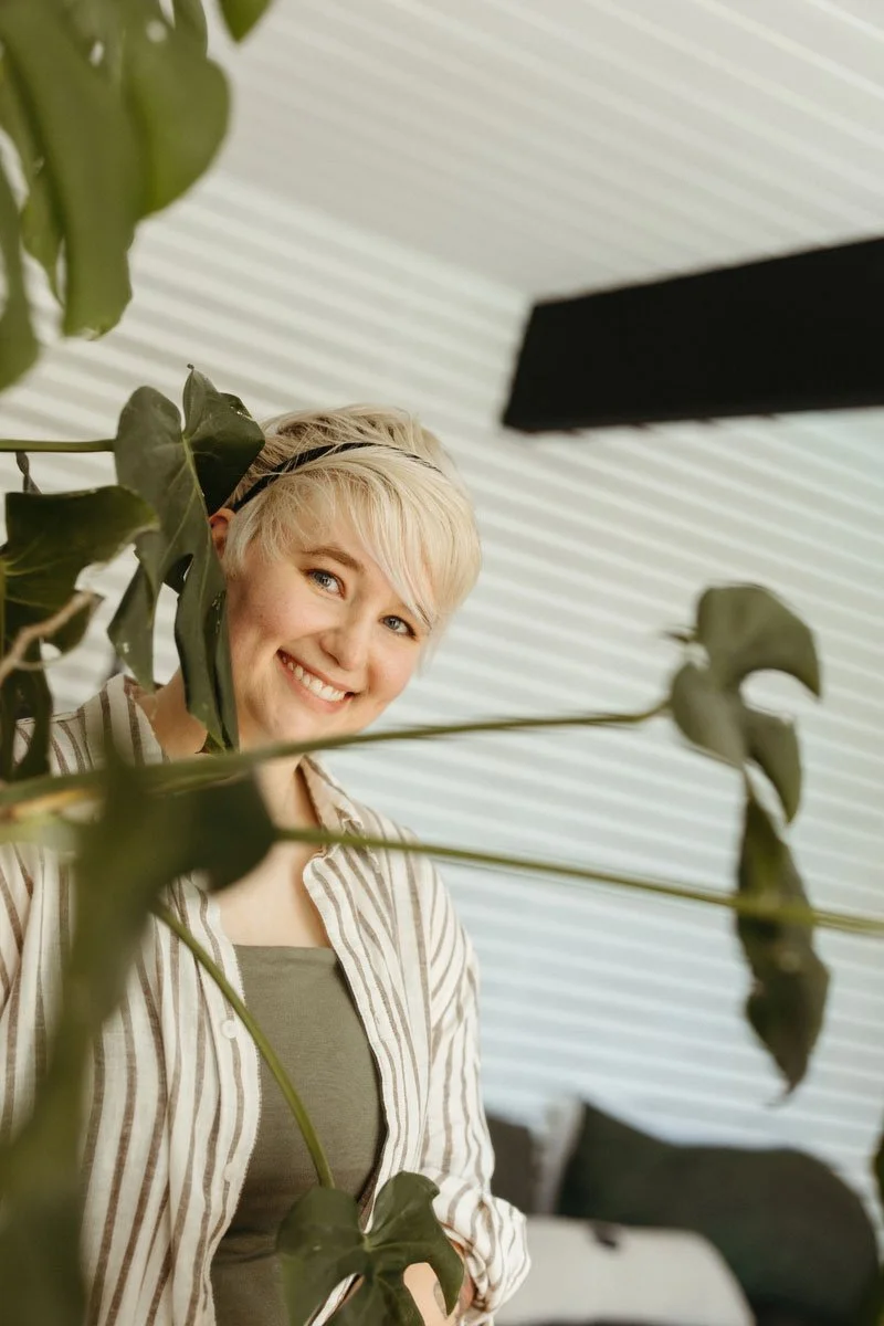 A woman with short blonde hair smiling behind large green plant leaves, indoors with white textured walls.