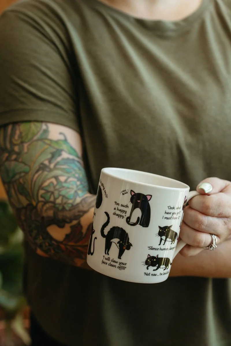 Lauren Abbott holding a white mug decorated with black cats, with a tattooed arm visible.