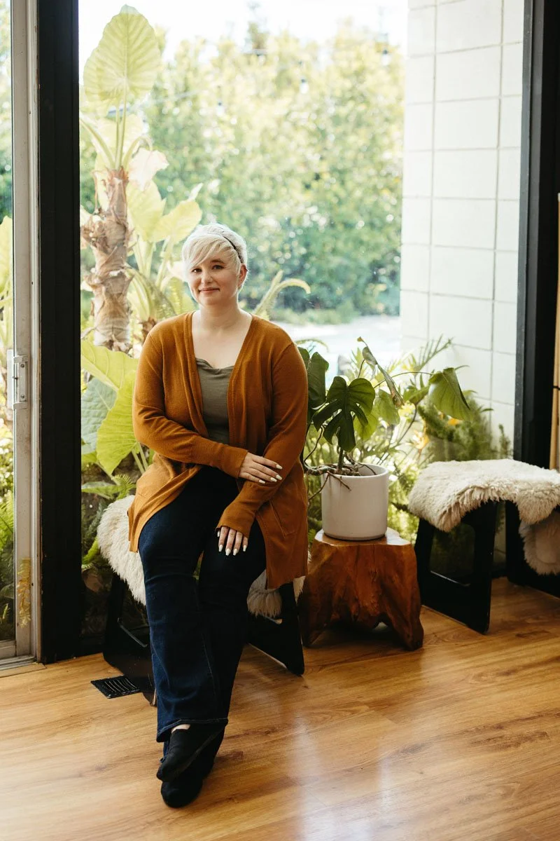 Lauren Abbott sitting on a bench inside near a large window, with green plants wearing a mustard-colored cardigan and black pants.