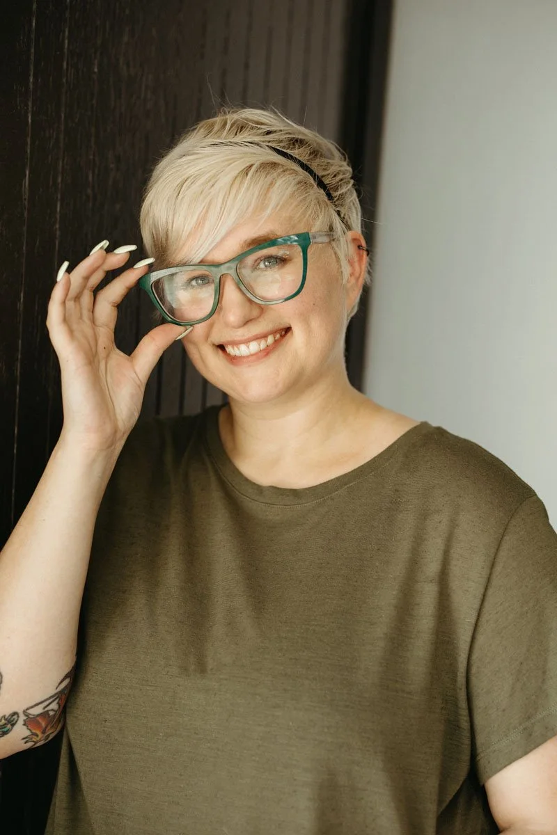 Lauren Abbott wearing glasses and a green T-shirt, smiling and holding her glasses with one hand.