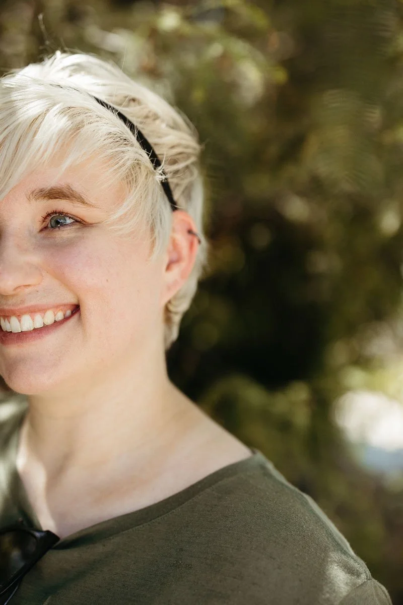 Close-up of a smiling woman with short blonde hair, wearing a black headband and green shirt, outdoors with greenery in the background.