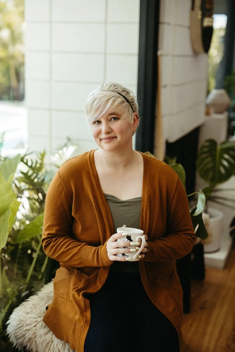 Lauren Abbott sitting indoors near a window, holding a mug and smiling, with green plants in the background.
