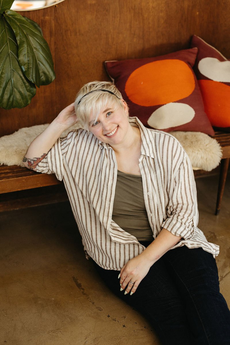 Lauren Abbott sitting on the floor next to a wooden bench with colorful pillows and a large green plant nearby.