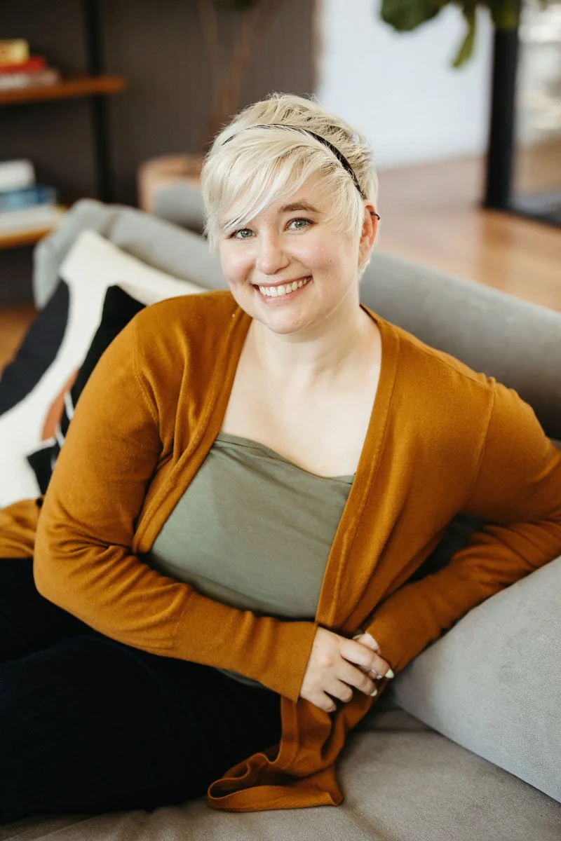Lauren Abbott, smiling and sitting on a gray couch in a living room.