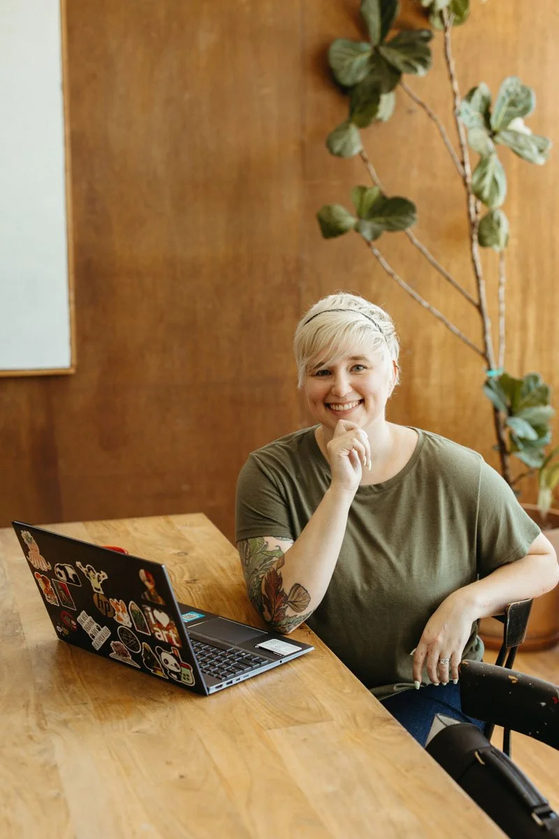 Lauren Abbott sitting at a wooden table with a laptop covered in colorful stickers. She is smiling and resting her chin on her hand in a relaxed pose.