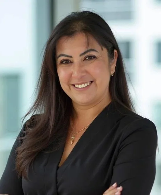 A smiling woman with long dark hair wearing a black blazer and jewelry, standing indoors with a blurred cityscape in the background.