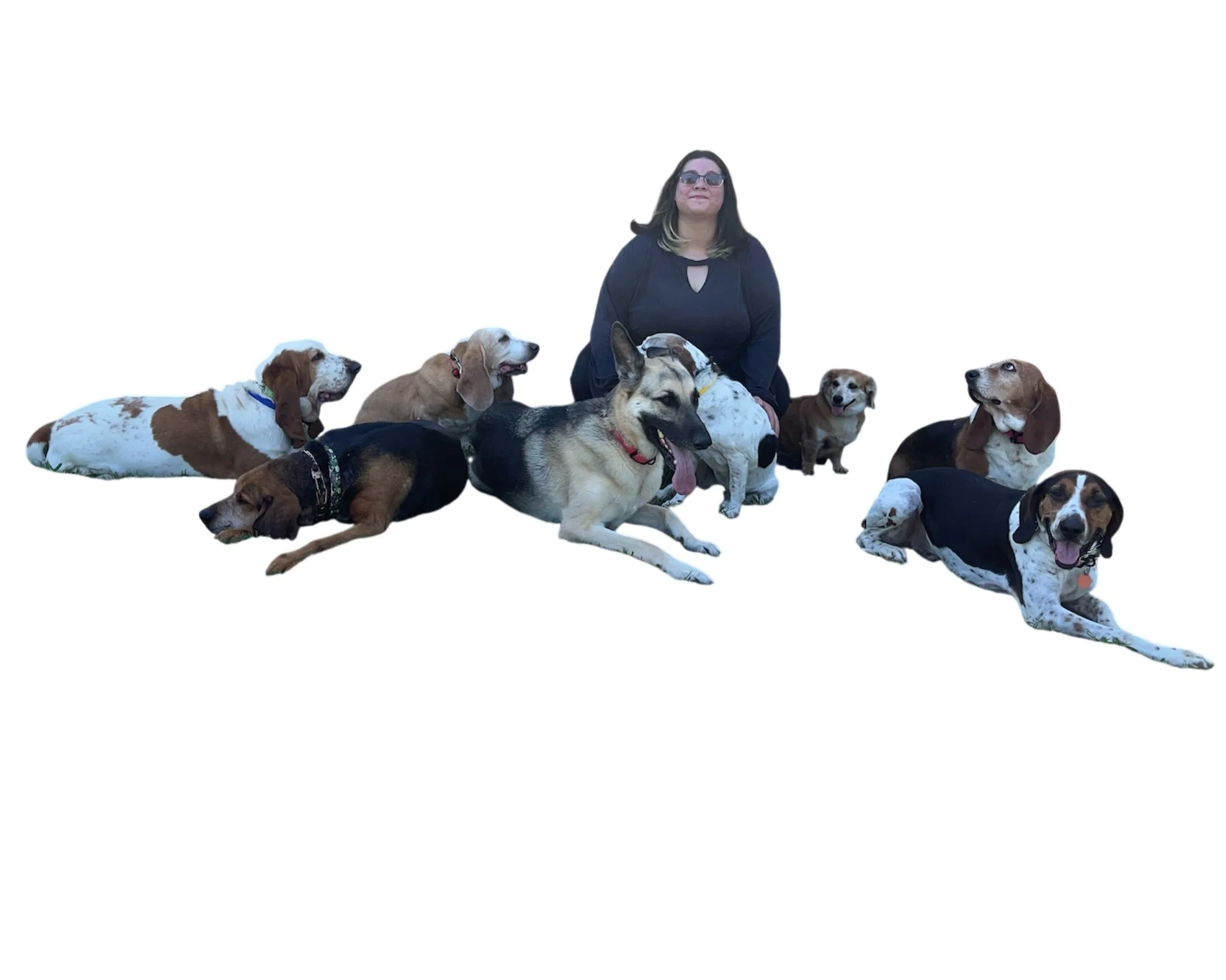 A woman with dark hair and sunglasses sitting on the ground surrounded by several dogs of various breeds and sizes.