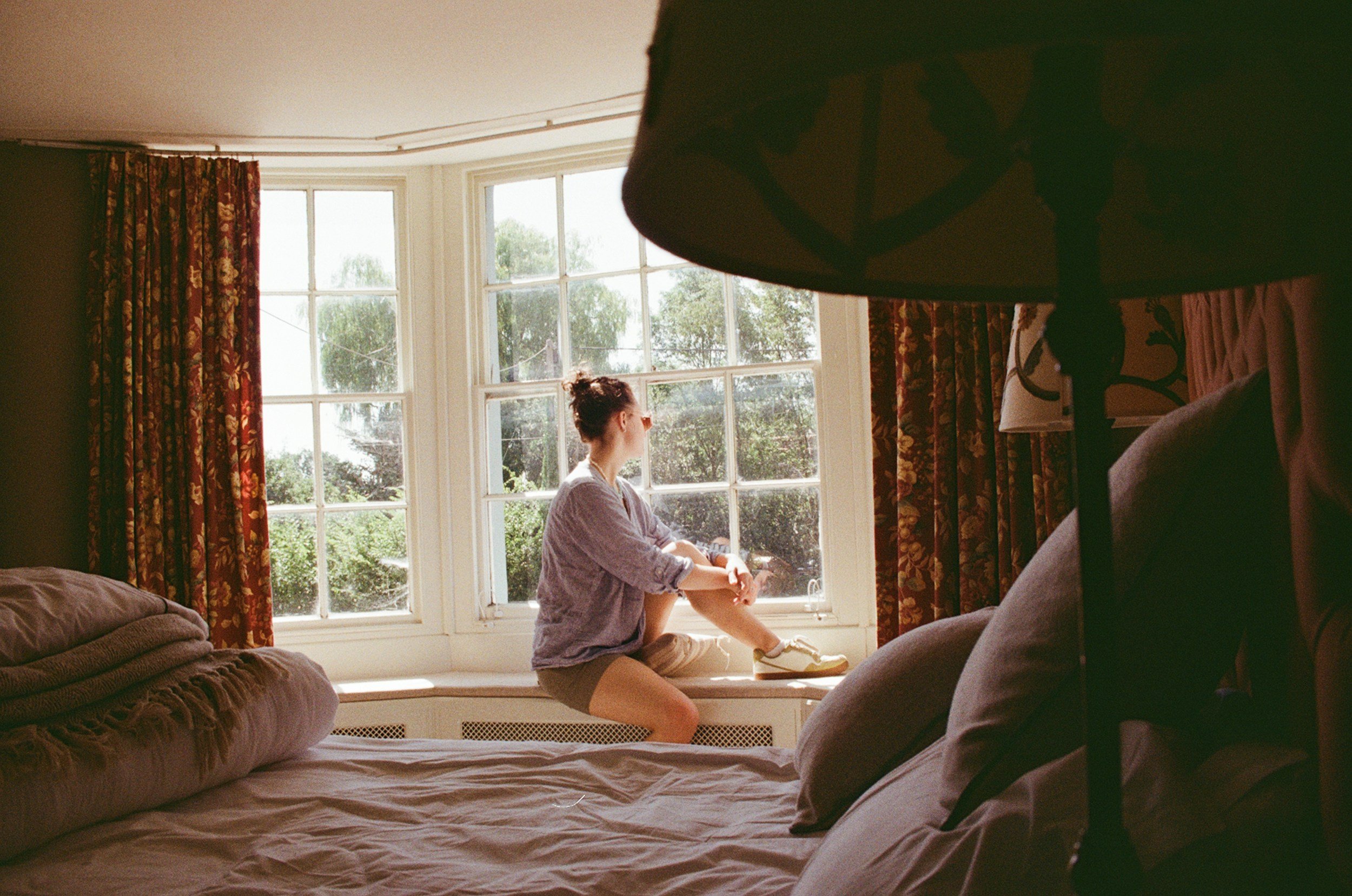 A woman sitting on a window ledge near a large window with floral curtains, looking outside at trees, with sunlight illuminating her and the room.