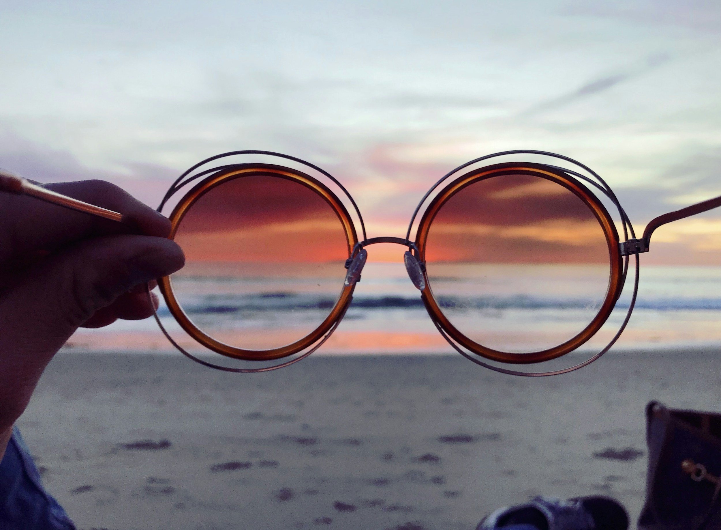 A person holding sunglasses in front of a beach at sunset, with the ocean, sandy shore, and colorful sky visible behind.