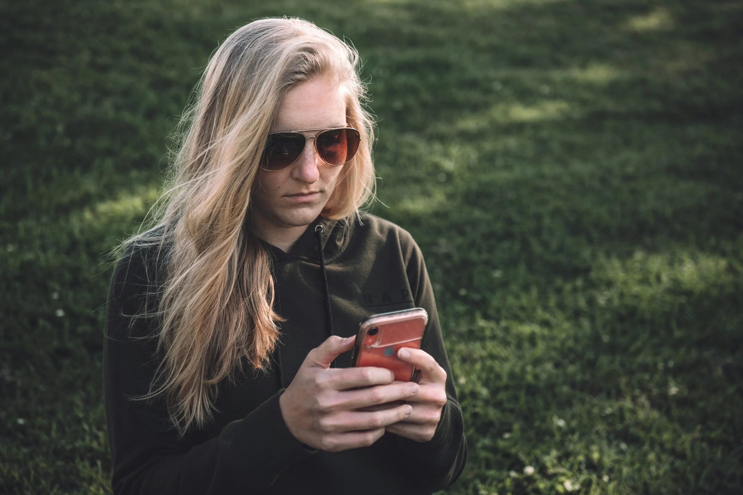 Young woman with long blonde hair wearing sunglasses and a dark hoodie, looking at her phone outdoors on a grassy area.