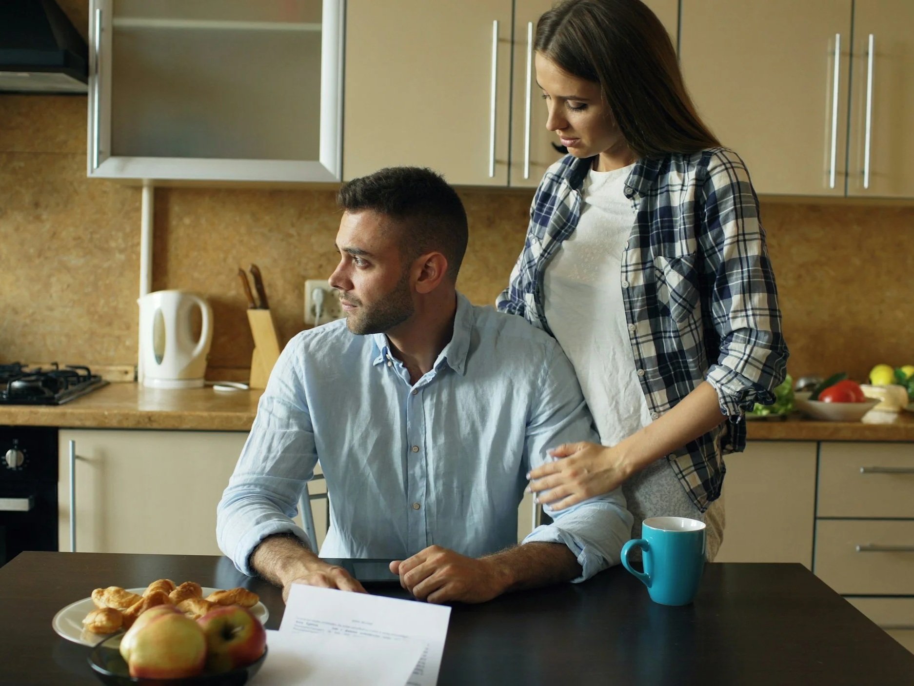 A woman standing behind a seated man at a kitchen table, looking down at a document with a cup of coffee and a plate of apples and pastries in the foreground, with kitchen cabinets and countertops in the background.