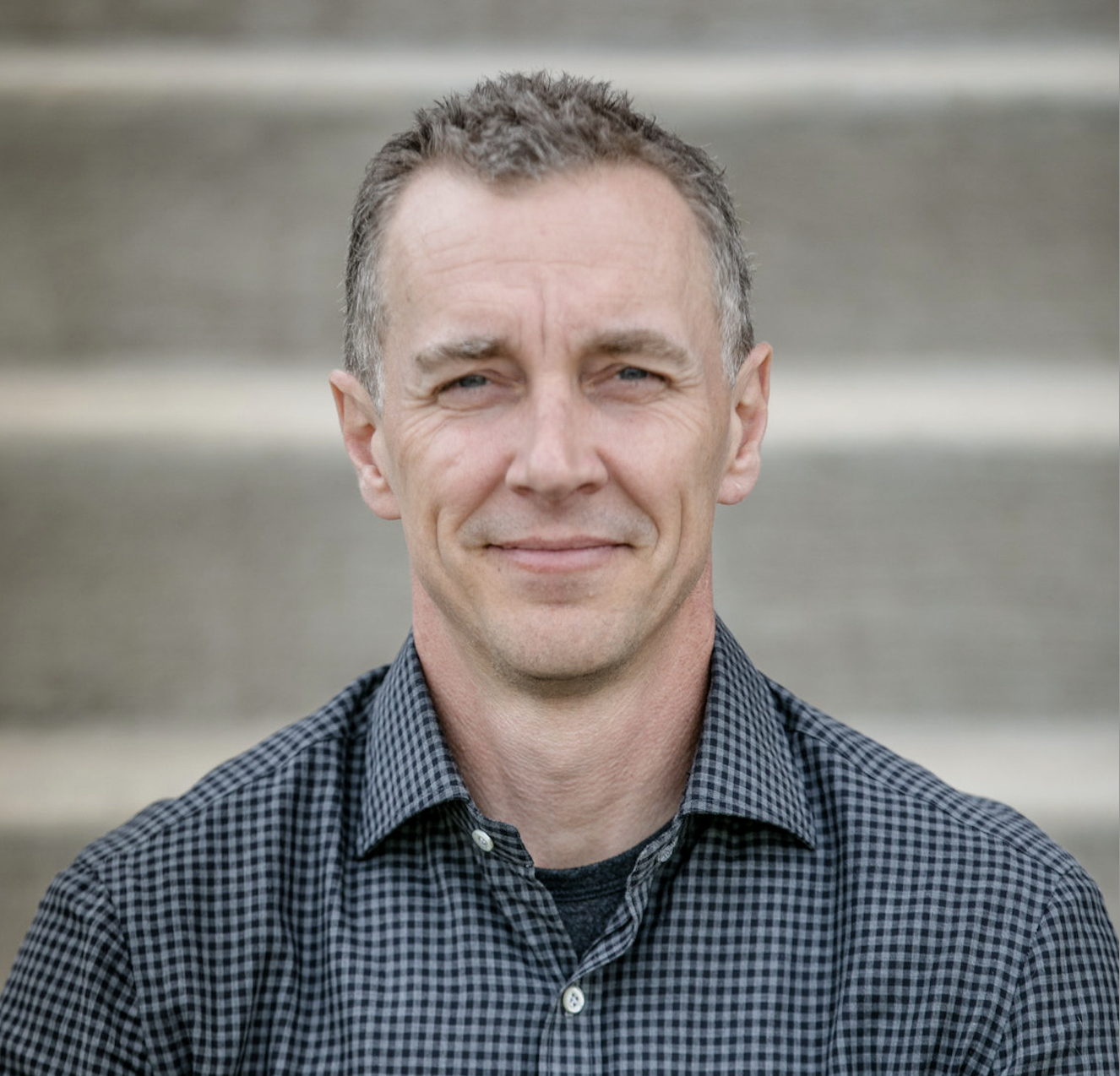 A middle-aged man with short gray hair, blue eyes, and light skin, smiling slightly, wearing a dark checkered shirt, standing outdoors on gray concrete stairs.