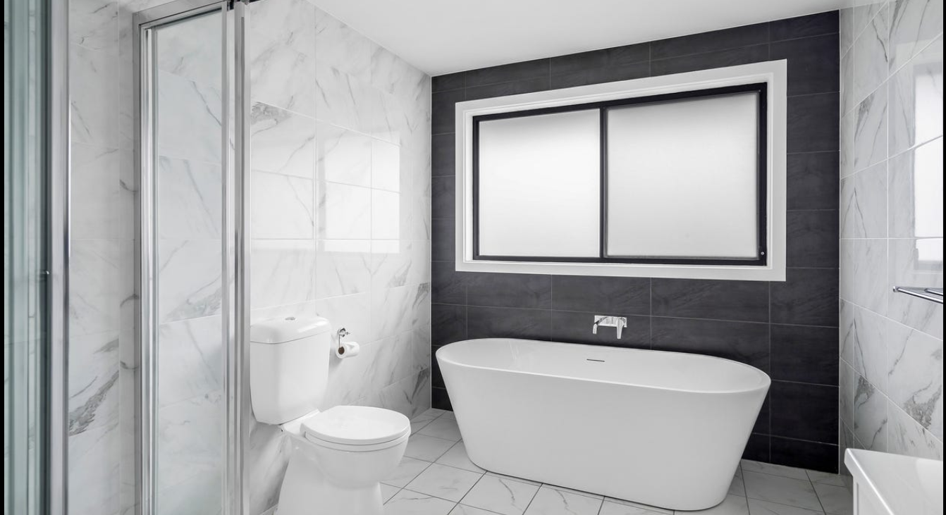 Modern bathroom with a white freestanding bathtub, a toilet, and tiling on the walls and floor, with a large frosted window.