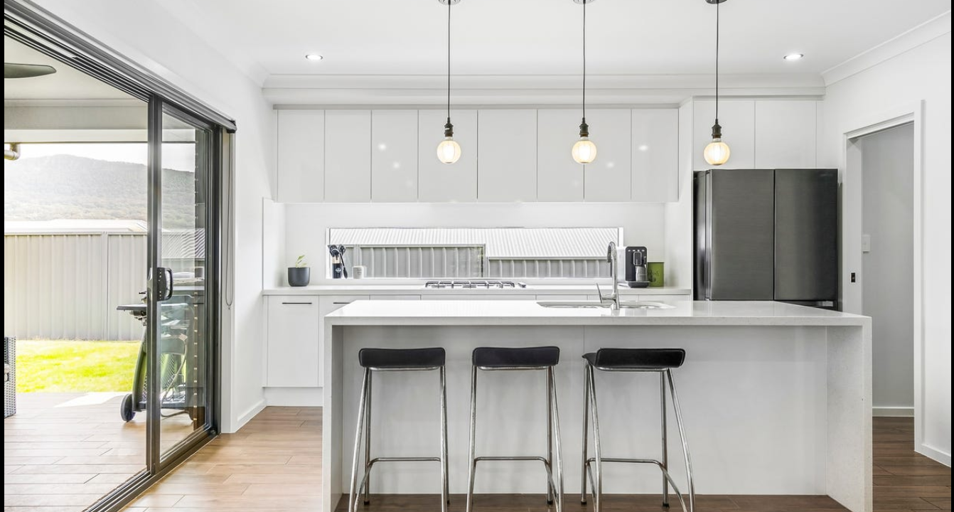 Modern white kitchen with a large island, three black bar stools, hanging light bulbs, a stainless steel refrigerator, a coffee maker, and a sliding glass door leading outside to a grassy yard.