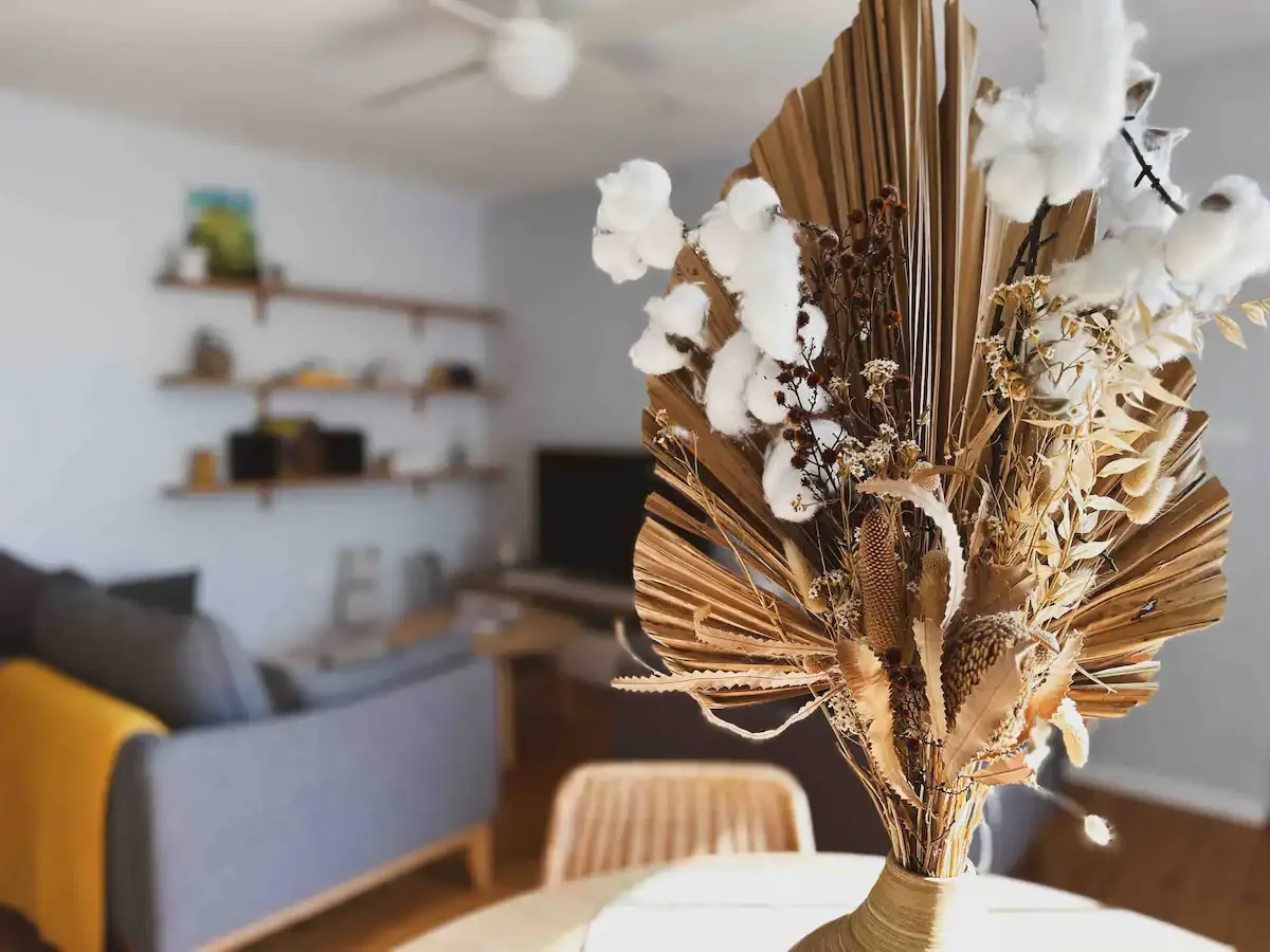 A dried flower arrangement with cotton, grasses, and leaves in a beige vase, placed on a table in a cozy living room.