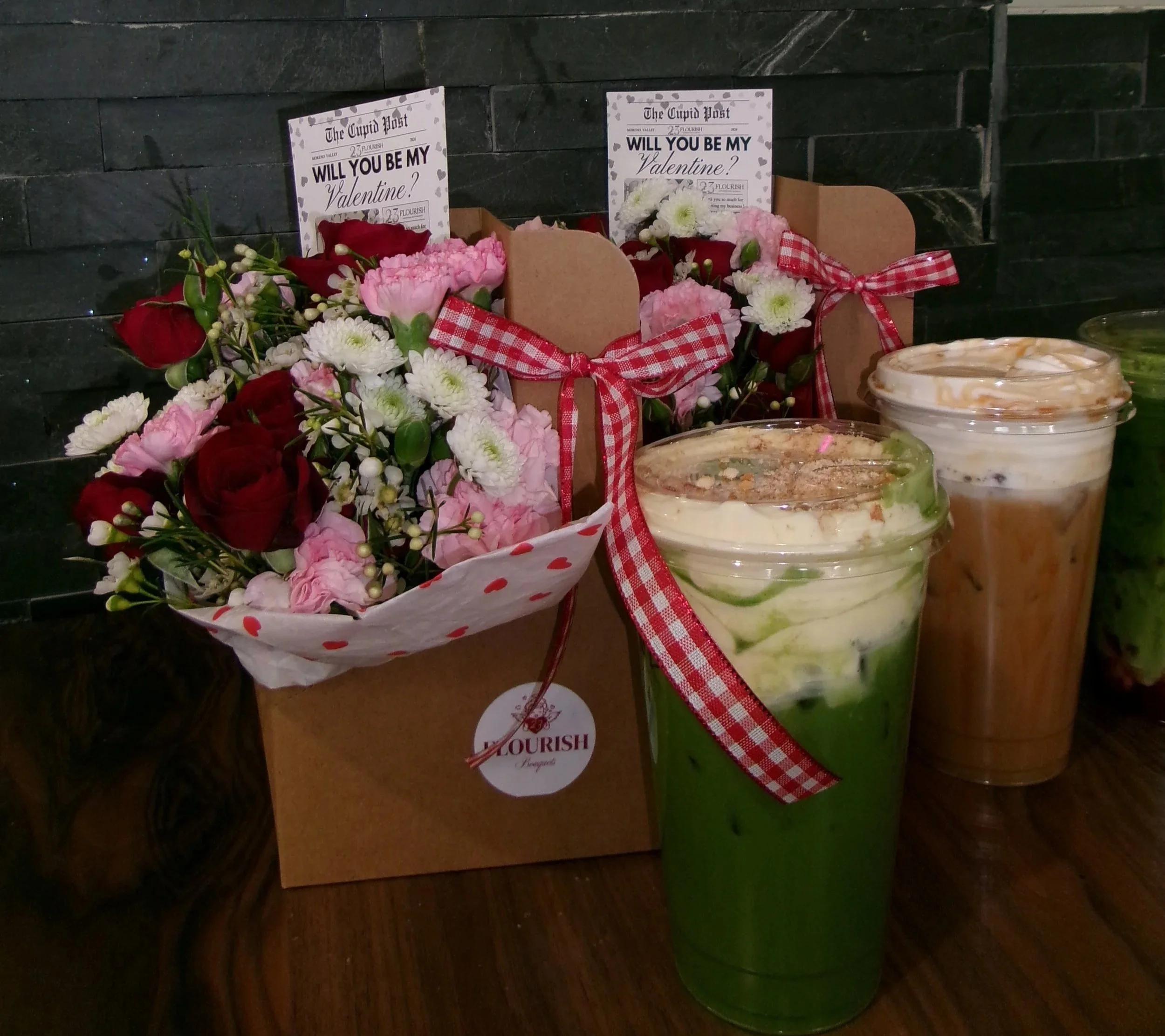A bouquet of red and pink flowers with green leaves and white filler flowers, decorated with a red and white gingham ribbon, placed next to two cups of a layered drink with green and white swirls, against a dark tiled wall.