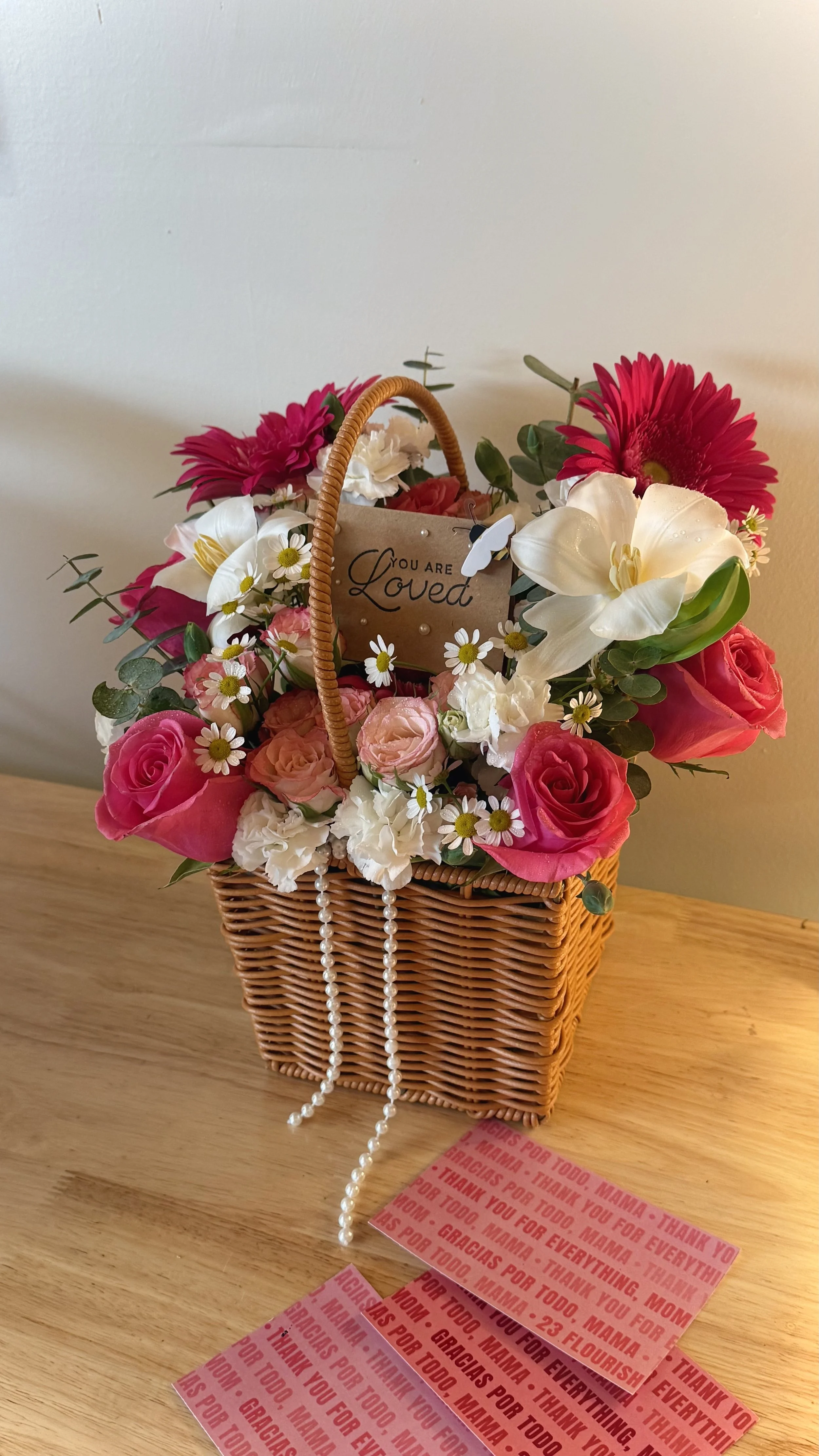 A wicker basket filled with various colorful flowers including pink roses, white tuberoses, red gerbera daisies, and small daisies, with a sign that reads 'You are Loved' attached to the basket. There are strings of pearl beads draped over the basket, and pink thank you cards with repeated words of gratitude are placed on the table.
